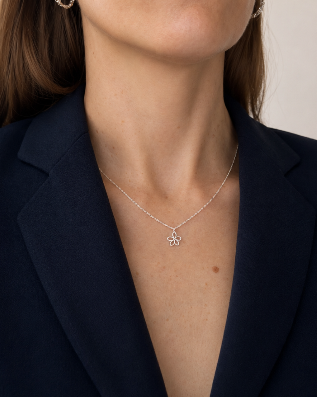 A close-up of a woman wearing a delicate silver necklace with a small flower-like pendant, styled with a dark navy blazer.